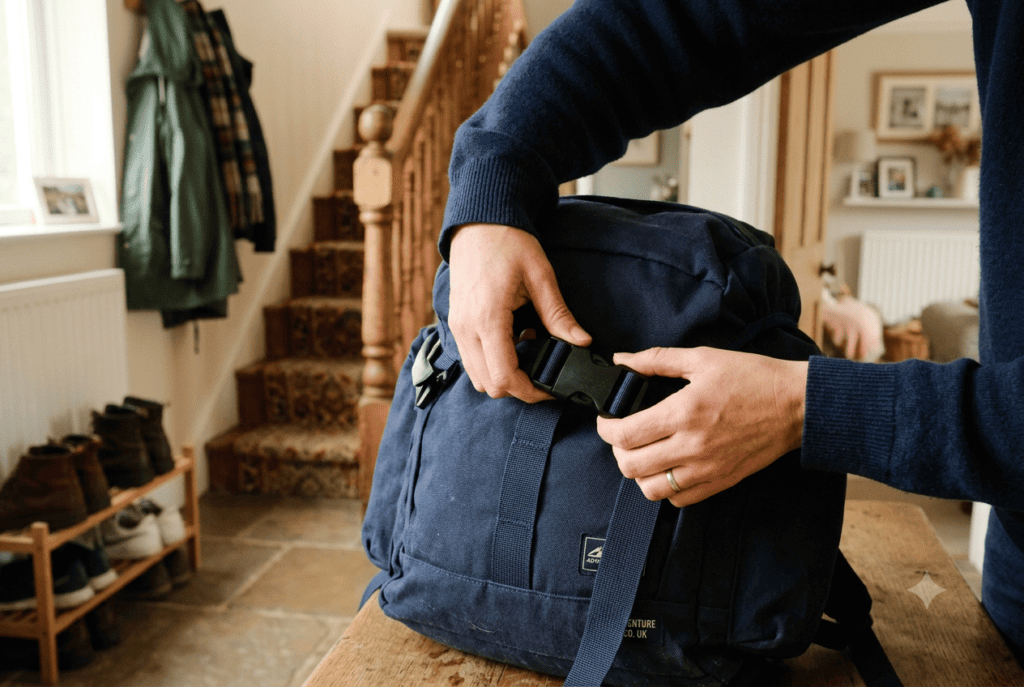A person fastening the buckle on a packed navy rucksack in a British hallway, ready to grab and go.